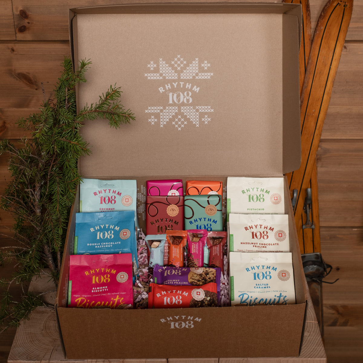 An open Rhythm 108 gift box filled with assorted packaged snacks and biscuits, set against a rustic wood background with pine branches and a ski boot prop.