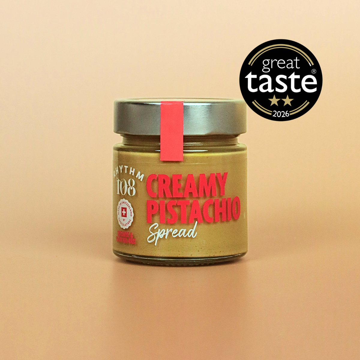 A glass jar of Rhythm 108 Creamy Pistachio Spread with a silver lid and a red seal, set against a solid beige background with a 'great taste 2026' award badge in the top right corner.