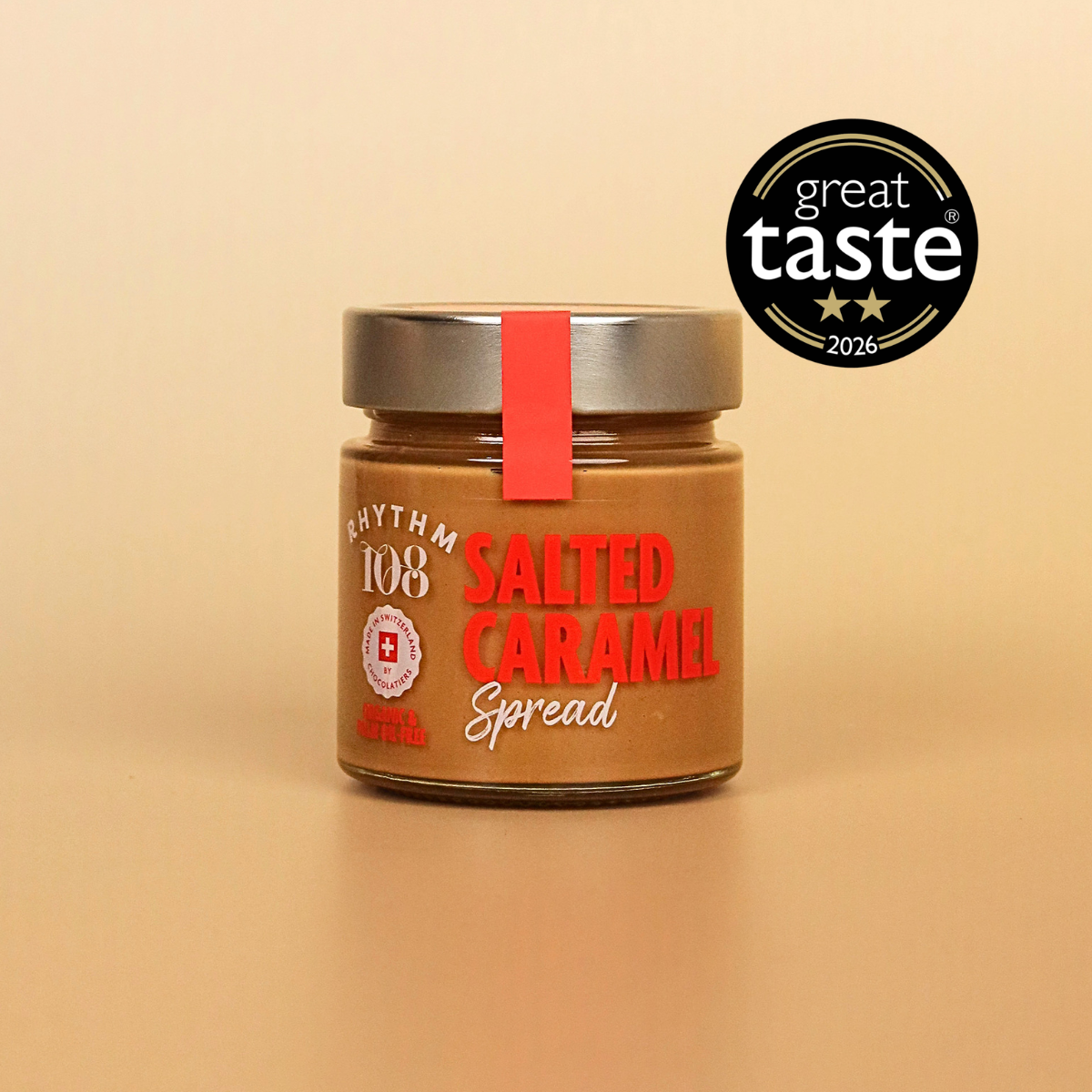 A jar of Rhythm 108 Salted Caramel Spread on a warm beige background, featuring a silver lid with a red ribbon and a black 'great taste 2026' award badge in the upper right.